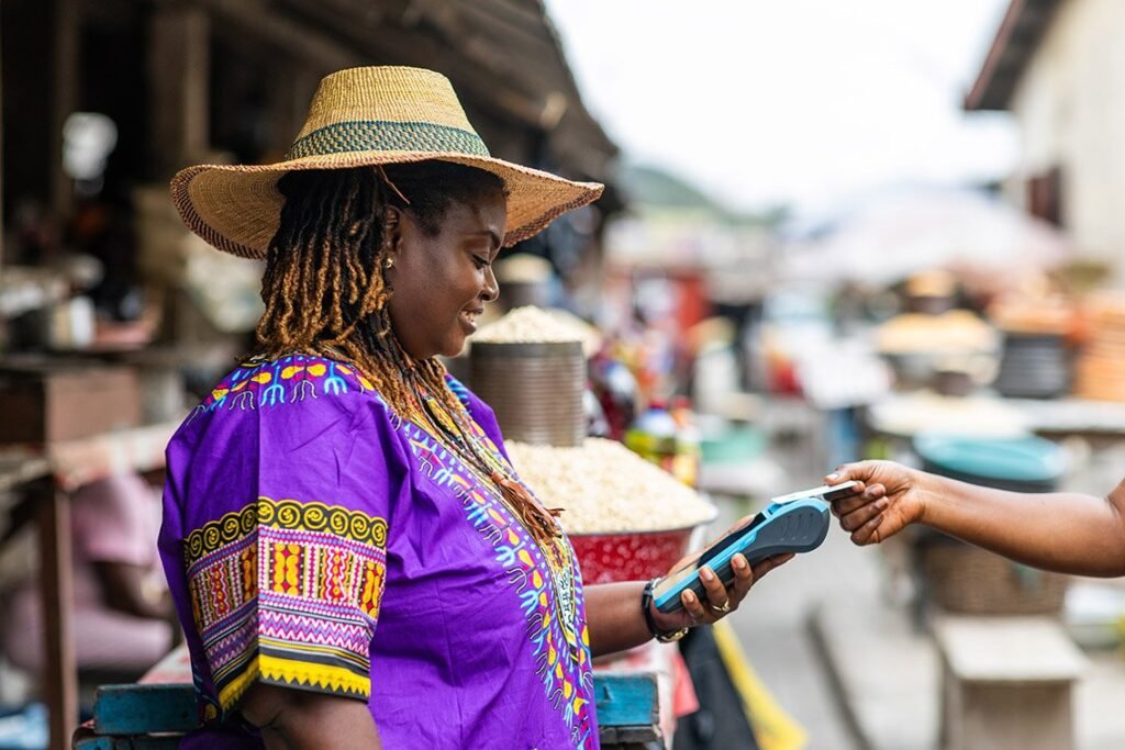 A smiling African woman in vibrant purple traditional attire with colorful patterns and a wide straw hat stands at an outdoor market stall. She holds a blue mobile payment terminal while accepting a card from a customer's hand, surrounded by market goods like grains and baskets in a bustling rural or semi-urban setting.