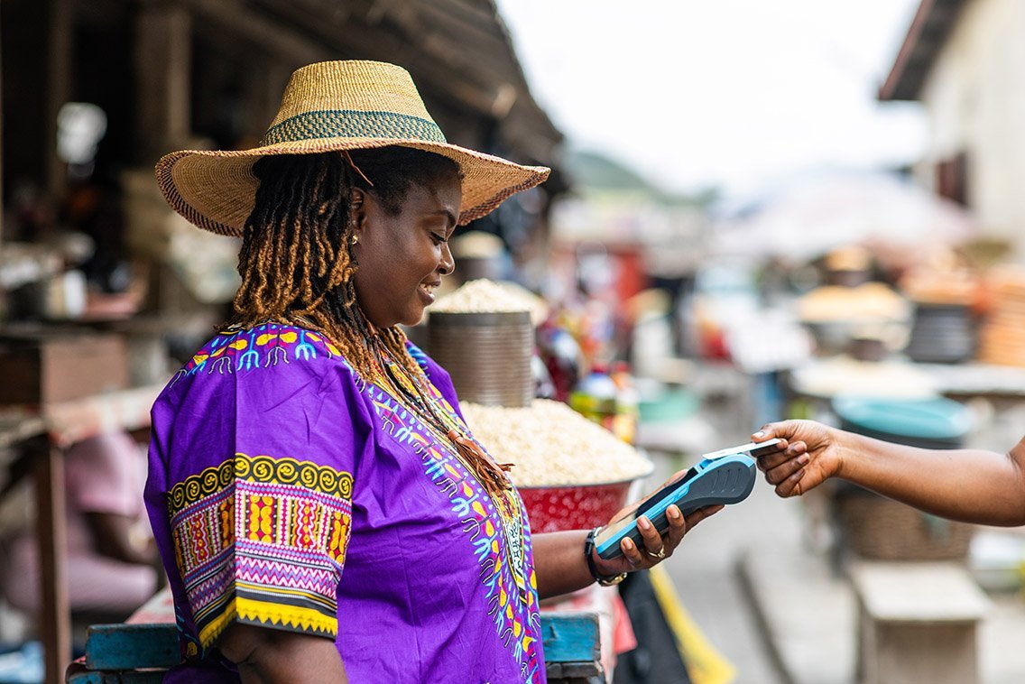 A smiling African woman in vibrant purple traditional attire with colorful patterns and a wide straw hat stands at an outdoor market stall. She holds a blue mobile payment terminal while accepting a card from a customer's hand, surrounded by market goods like grains and baskets in a bustling rural or semi-urban setting.