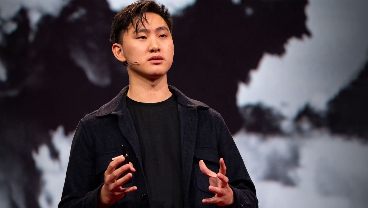 Un très jeune homme asiatique, portant une veste noire et un micro-casque, parle avec passion sur scène devant un grand fond représentant une carte du monde stylisée en sombre.