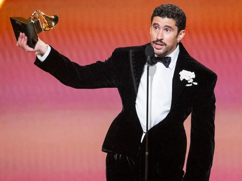 Un homme aux cheveux bouclés, moustachu, en smoking de velours noir avec nœud papillon et fleur blanche à la boutonnière, brandit un trophée Grammy sur scène devant un fond rose-orange.